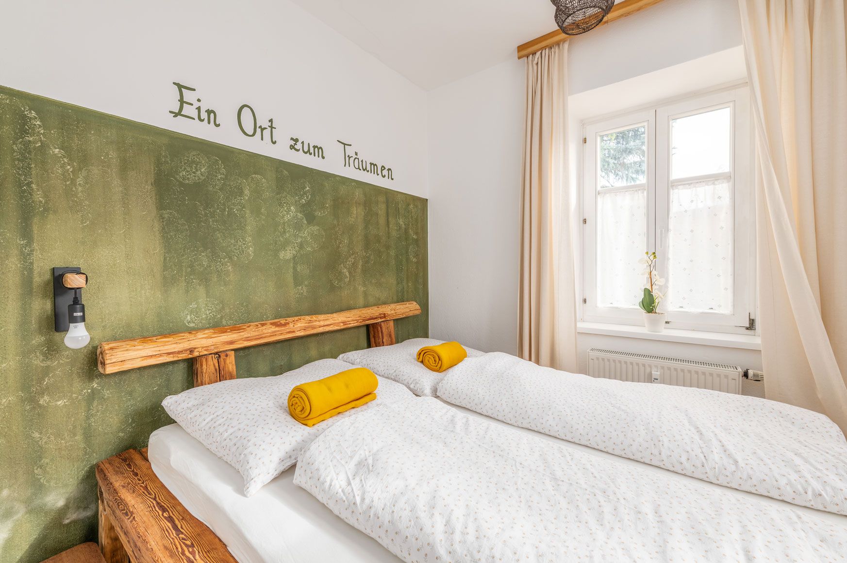 Schlafzimmer der Ferienwohnung mit Doppelbett, Holzrahmen, grüner Wand mit Schrift 'Ein Ort zum Träumen' und gelben Handtüchern auf weißen Bettdecken